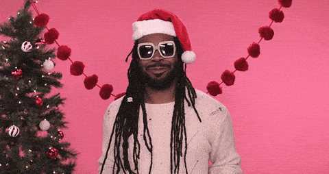 Decorações de Natal em espanhol chapeu-de-papai-noel-em-espanhol
DESCRIÇÃO DA IMAGEM: Um homem de dreadlocks e óculos escuros dourados usa um gorro vermelho de Papai Noel e manda um beijo para a câmera usando as duas mãos. Ele está usando um suéter branco e, no fundo, vemos uma árvore de Natal e alguns pompons vermelhos unidos por um fio e pendurados como uma faixa.